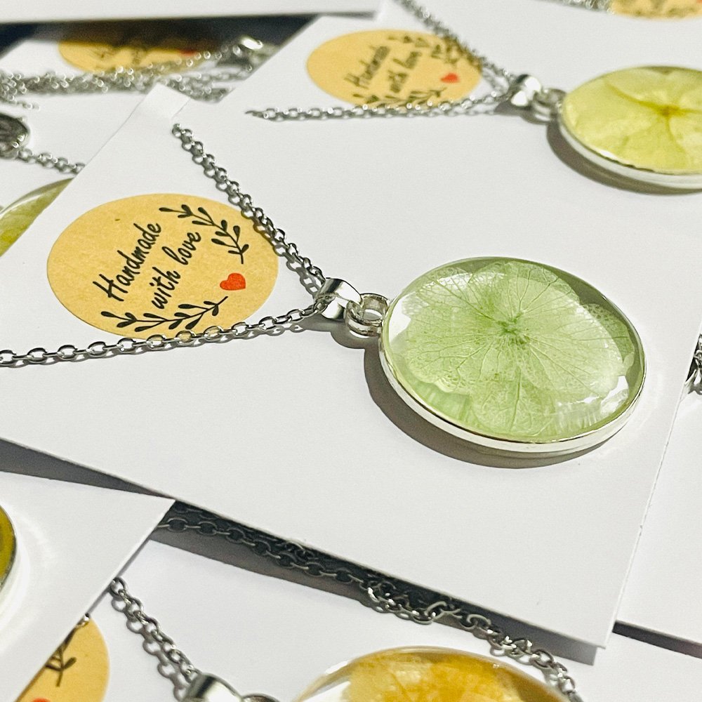 Handmade resin pendants with real green and yellow preserved flowers on silver chains, displayed on handmade packaging.